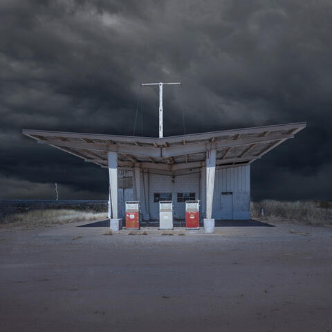Vintage gas station with distinctive angular roof canopy and red fuel pumps set against dramatic cloudy sky
